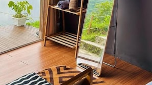 A room with a large mirror reflecting a scenic view, a wooden swing chair, a small shelf with a rolled towel, and a potted plant by the window. - Fortune Glamping (Garung)