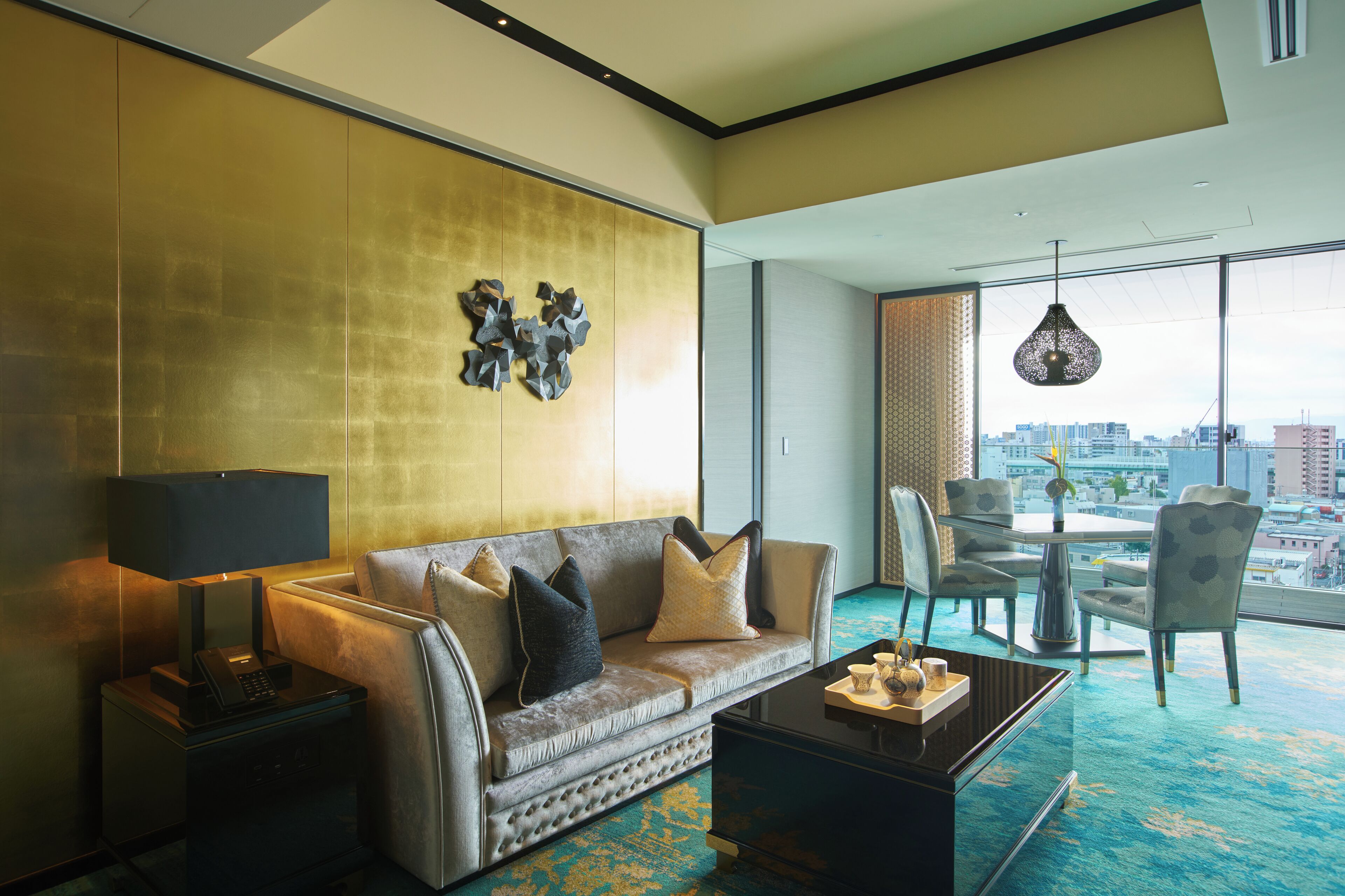 A modern hotel room with a sofa, armchairs, a coffee table, and a city view through large windows.