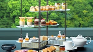 A three-tiered dessert stand with various pastries and desserts, a teapot, cups, and a bowl on a table with a view of a traditional Japanese castle in the background. - ESPACIO NAGOYA CASTLE (Nagoya)