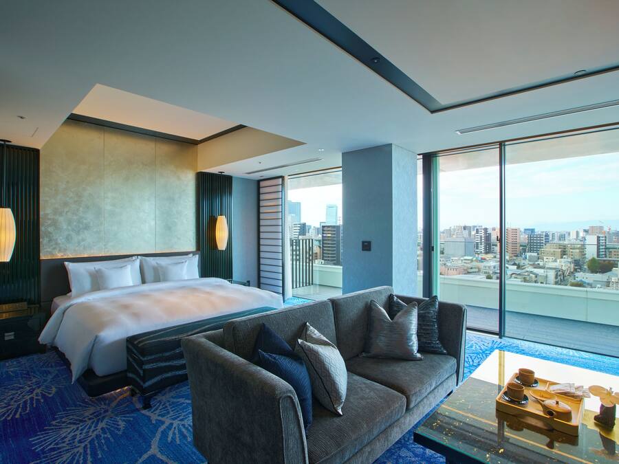 A modern hotel room with a large bed, a sofa, a glass coffee table, and a city view.