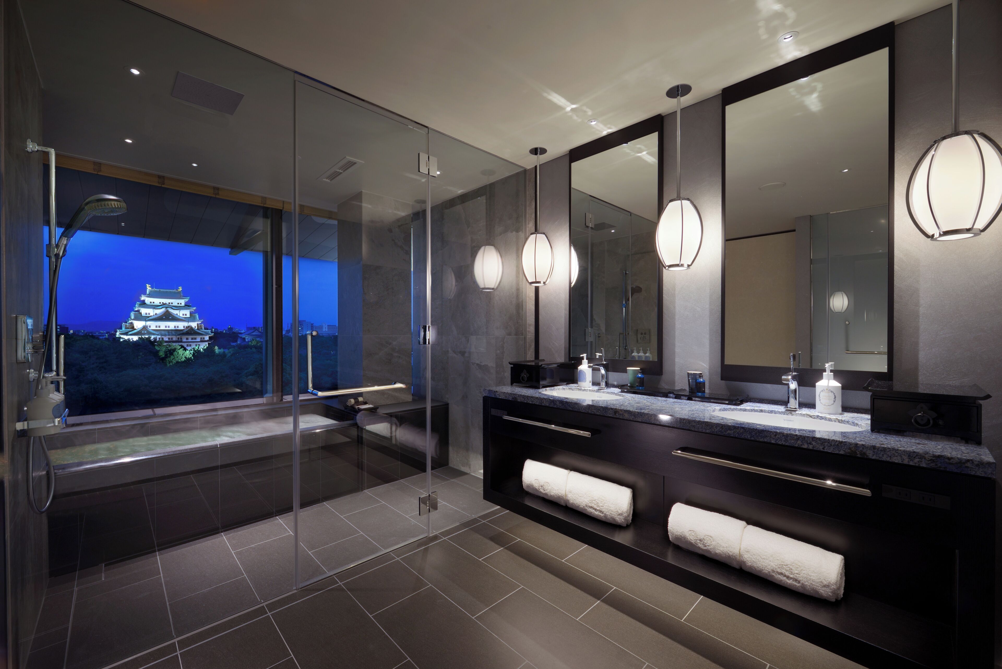 A modern bathroom with a large mirror, dual sinks, a glass-enclosed shower, and a view of a traditional building at night.