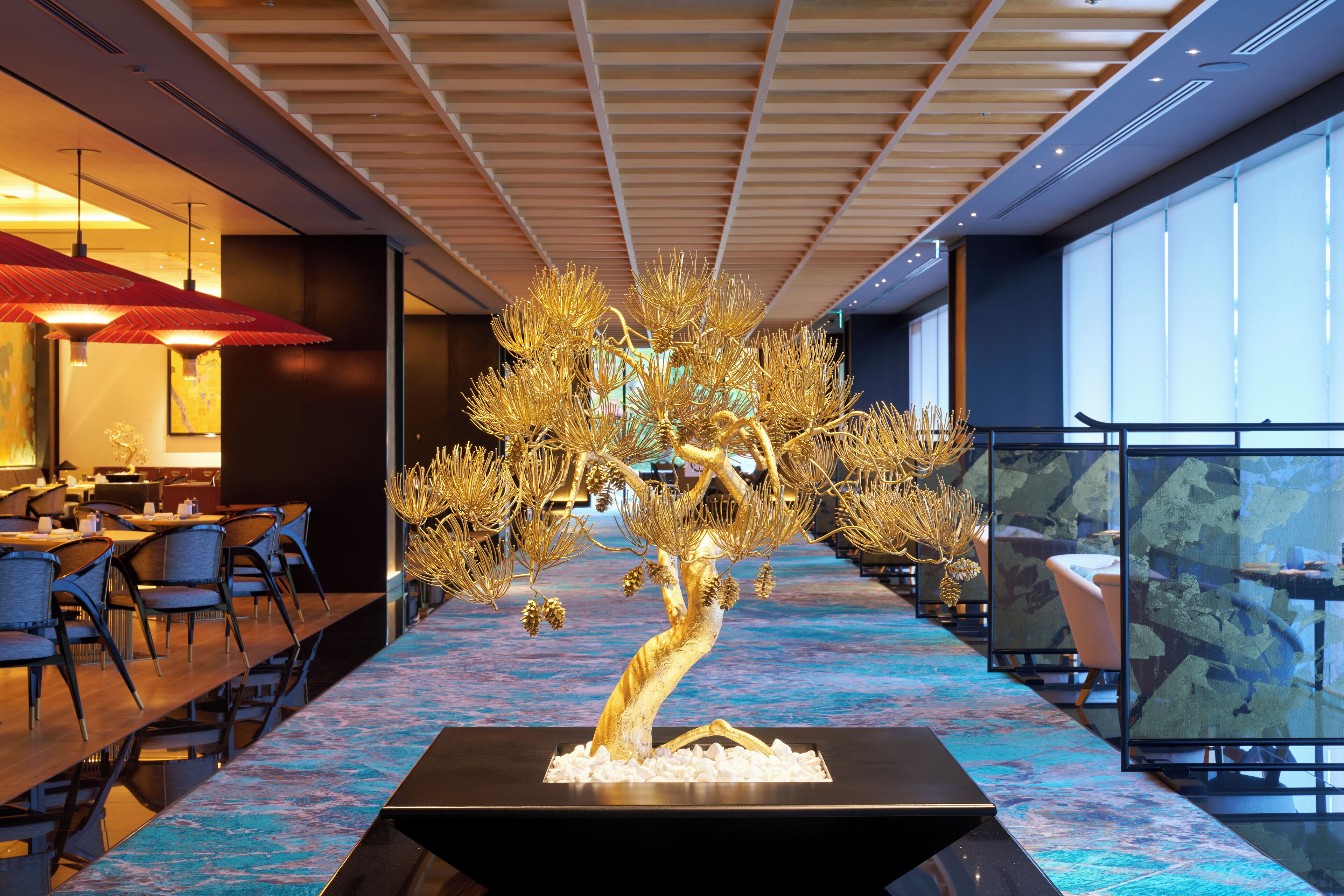 A modern hotel lobby with a central golden tree sculpture, dining area with tables and chairs, and large windows offering views.