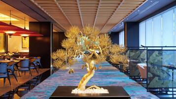 A modern hotel lobby with a central golden tree sculpture, dining area with tables and chairs, and large windows offering views.