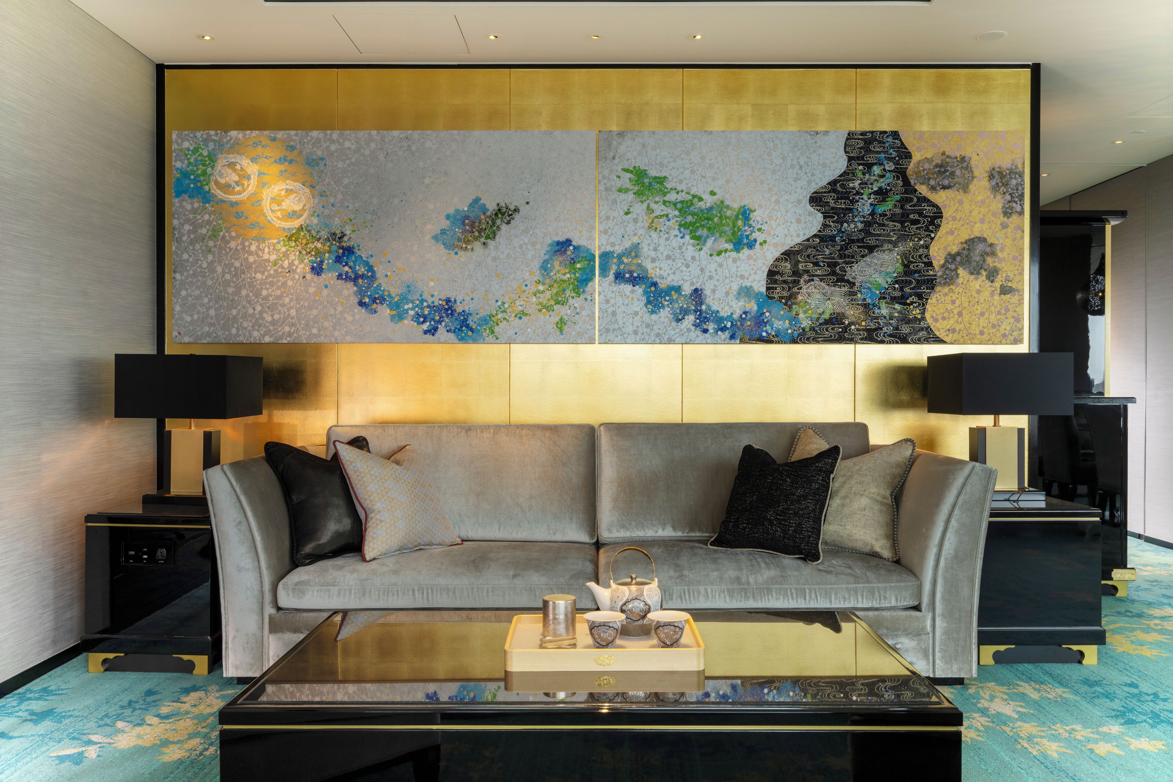 Una sala de estar moderna con un sofá gris, dos mesitas de noche con lámparas, una mesa de centro con objetos decorativos y un cuadro abstracto grande en la pared.