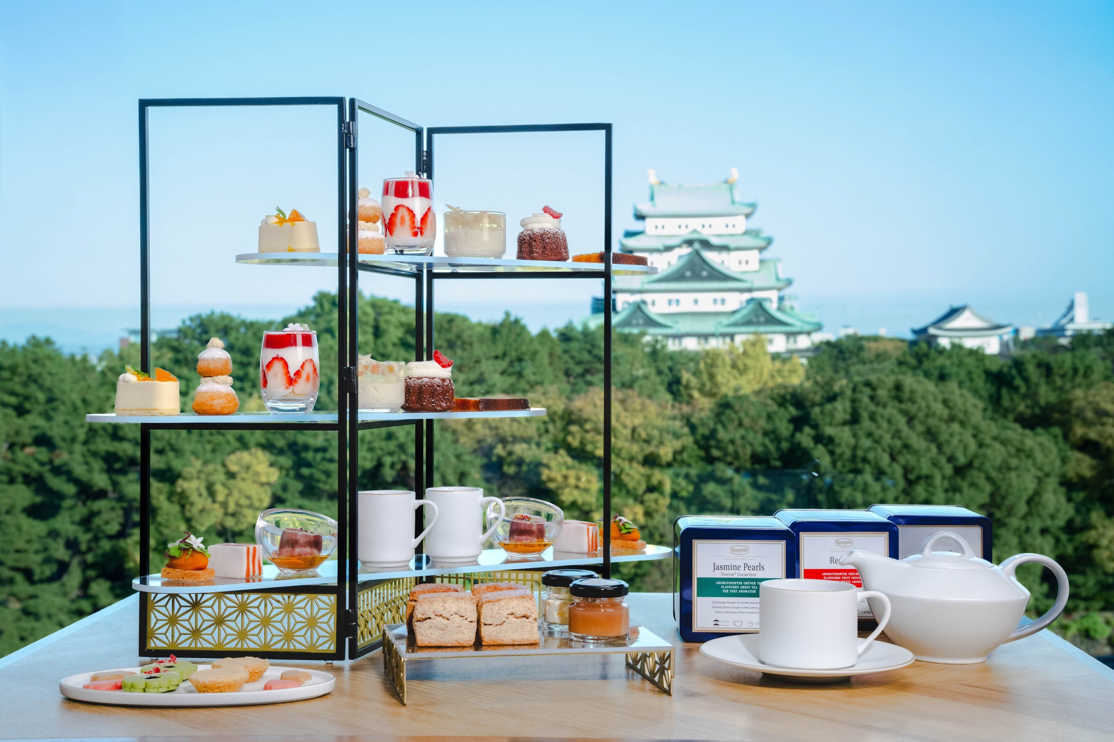 A tiered dessert display with various pastries and beverages, set against a scenic view of traditional architecture and lush greenery.
