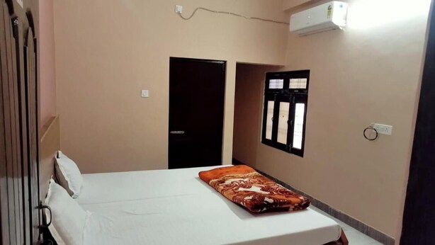 A single bed with a patterned blanket, a wall-mounted air conditioner, and a window with blinds. - Sankatha Guest House (VARANASI)