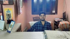 A man sitting at a counter in a room with a window, a sign indicating a check-out time of 12:00 noon, and a QR code on a card. - Sankatha Guest House (VARANASI)