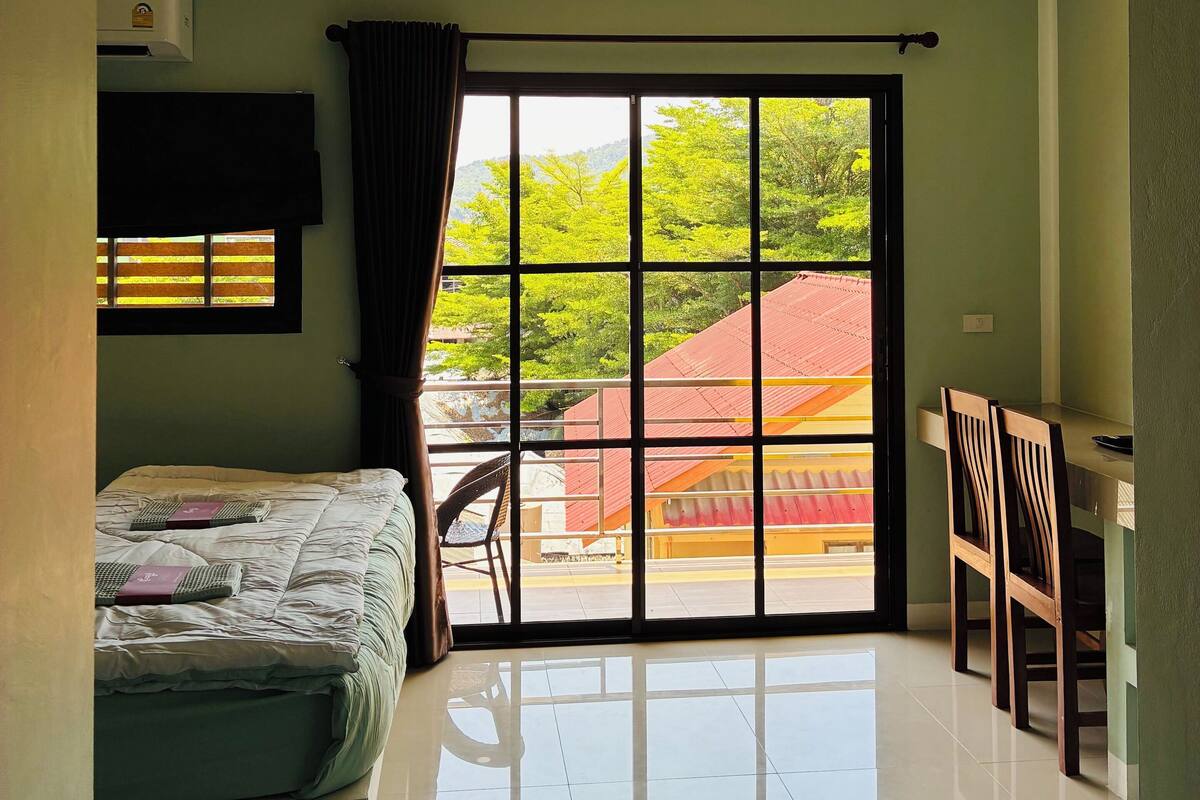 Ein Zimmer mit einem Bett, einem Sessel, einem Schreibtisch und einem großen Fenster mit Blick auf Grünflächen und Gebäude.