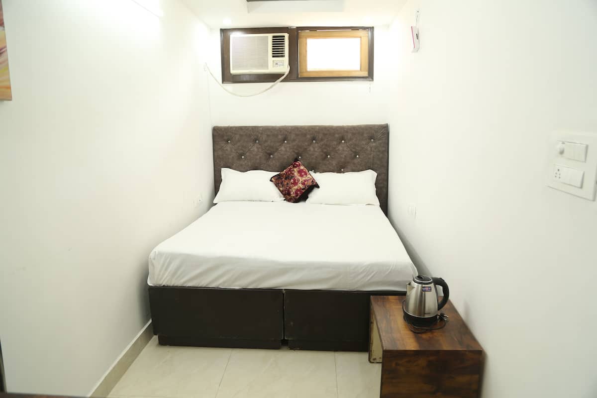 Una cama bien hecha con ropa blanca y una almohada decorativa, una mesita de noche con una tetera y una ventana con persianas.