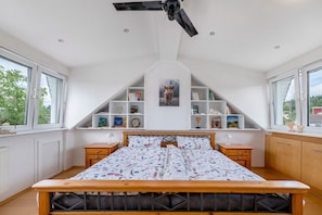 Quarto com teto inclinado, cama com colcha florida, ventilador de teto e estantes embutidas com livros e objetos decorativos.
