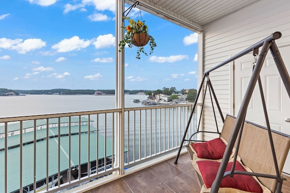 A balcony with a swing chair, a hanging plant, and a view of a river with boats.