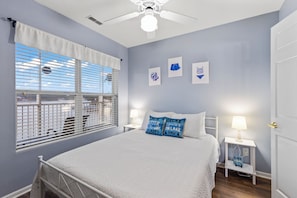 A bedroom with a bed, a nightstand with a lamp, a window with blinds, and wall art.