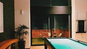 Una mesa de billar en una habitación con un ventanal grande, una planta en maceta y una pared de ladrillo visible a través del ventanal.
