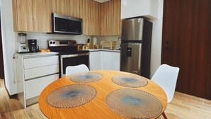 Una cocina compacta con armarios de madera, un microondas, una estufa y un refrigerador. Hay una mesa redonda con un mantel tejido y dos sillas blancas.