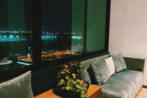 Una habitación con un ventanal grande que da a una vista nocturna de la ciudad. Hay un sofá con cojines y una mesita con una planta.