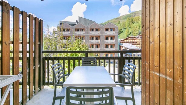 A balcony with a table and chairs, overlooking a building with multiple balconies and a mountainous landscape.