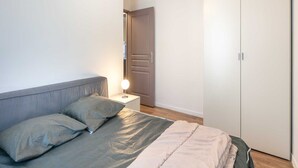 A bedroom with a bed, pillows, a bedside lamp, a wardrobe, and a door leading to another room.