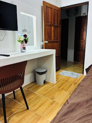 Une pièce avec un plancher en bois, un bureau blanc avec une chaise, une télévision, une fenêtre ornée d’un vase de fleurs et une porte portant le numéro 26.
