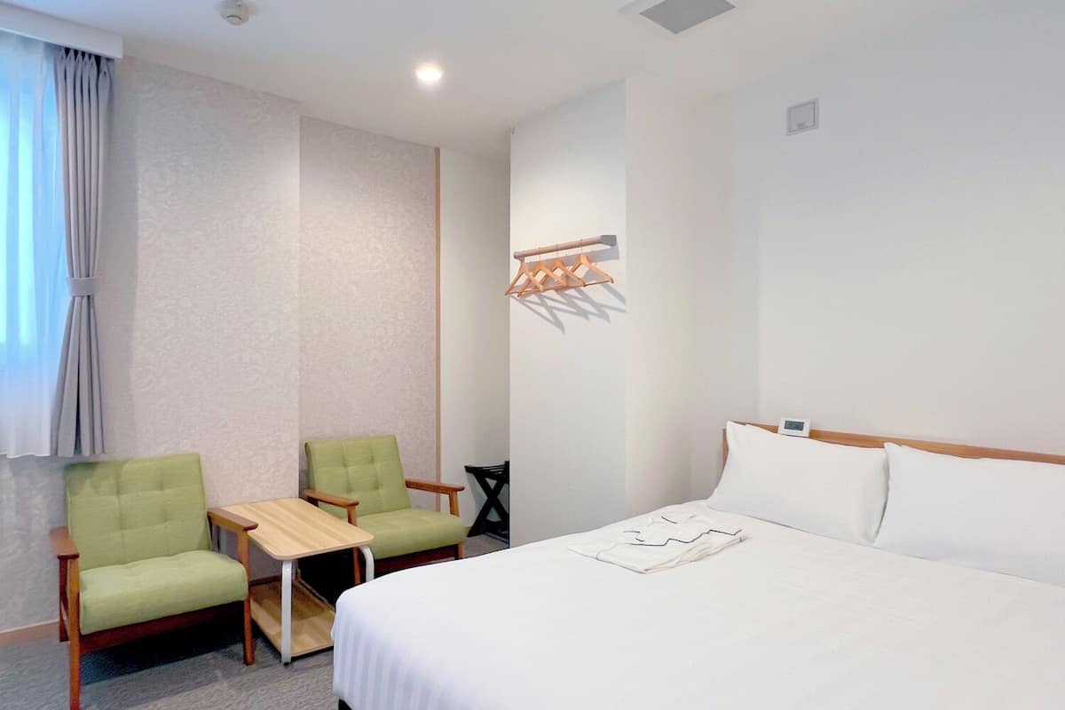 Una habitación de hotel con una cama, dos sillones verdes, una mesita y un perchero en la pared.