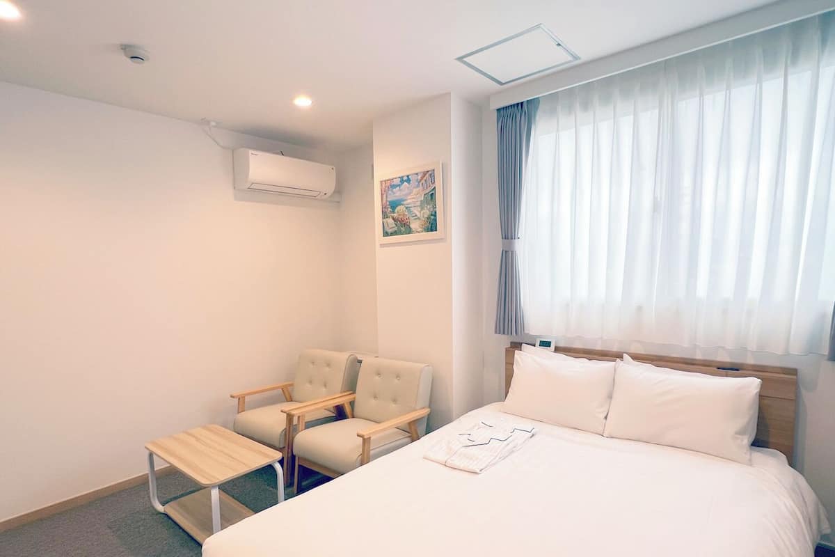Habitación de hotel con cama, dos sillones, una mesita, aire acondicionado, un cuadro en la pared y una ventana con cortinas.