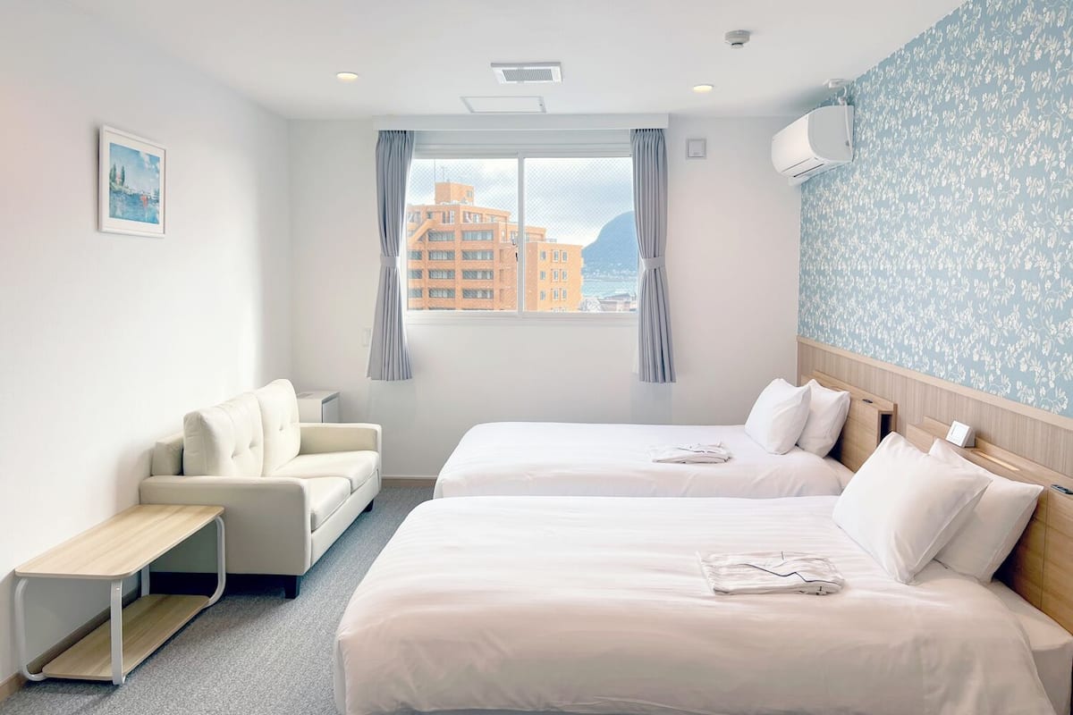 Habitación de hotel con dos camas, un sofá, una mesita y ventana con vista a edificios.