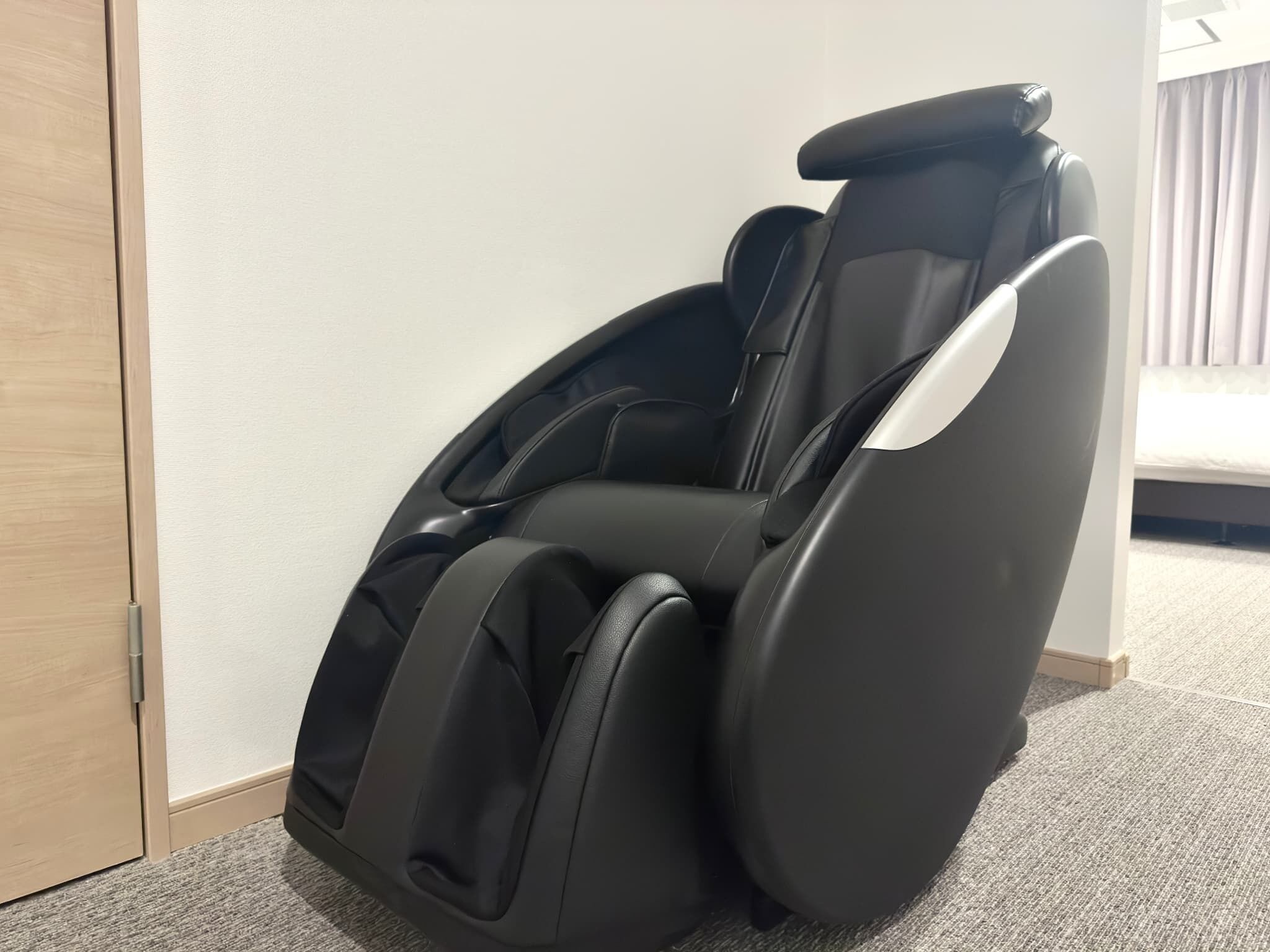 A massage chair in a room with a carpeted floor and a bed in the background.