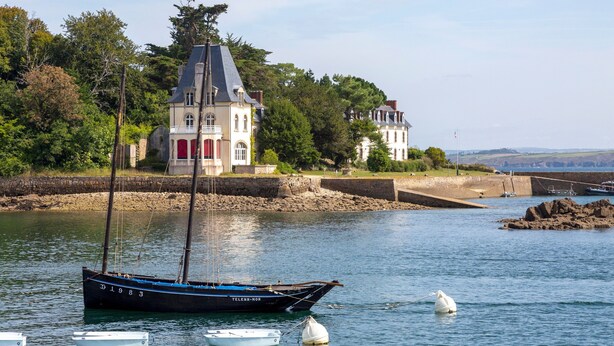 Eine Küstenlandschaft mit einem Haus, einem Segelboot und einem Bojen im Wasser.