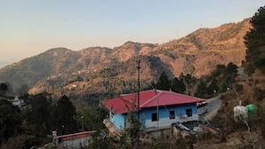 A rural landscape with a single house featuring a red roof, surrounded by hills and sparse vegetation. - Sumangalam Homestay (Pauri)