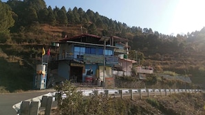 A two-story building with a signboard, located near a road with a guardrail. - Sumangalam Homestay (Pauri)