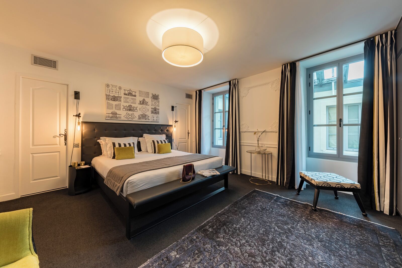 A modern hotel room with a large bed, bedside tables, a small bench, and a view of the city through large windows.
