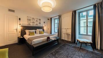 A modern hotel room with a large bed, bedside tables, a small bench, and a view of the city through large windows.