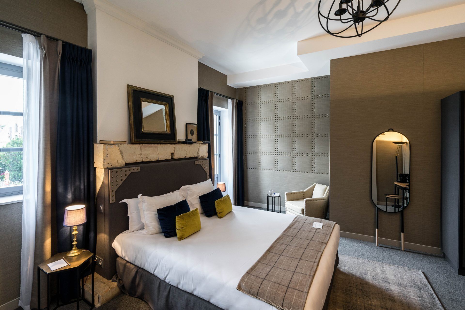 Quarto de hotel com uma cama grande, mesa de cabeceira com abajur, uma cadeira, espelho e painel texturizado na parede.