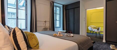 A hotel room with a large bed, a small table with a tray of fruit and a bottle, and a chair in the corner.