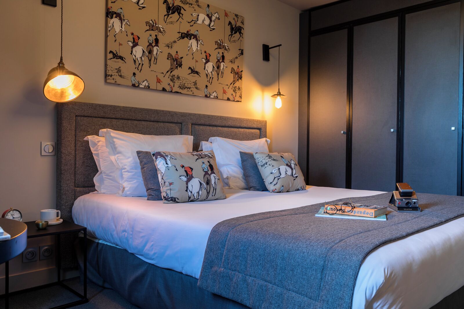 A neatly made bed with patterned pillows, a bedside table with a cup and a book, and a wall with a horse-themed artwork.