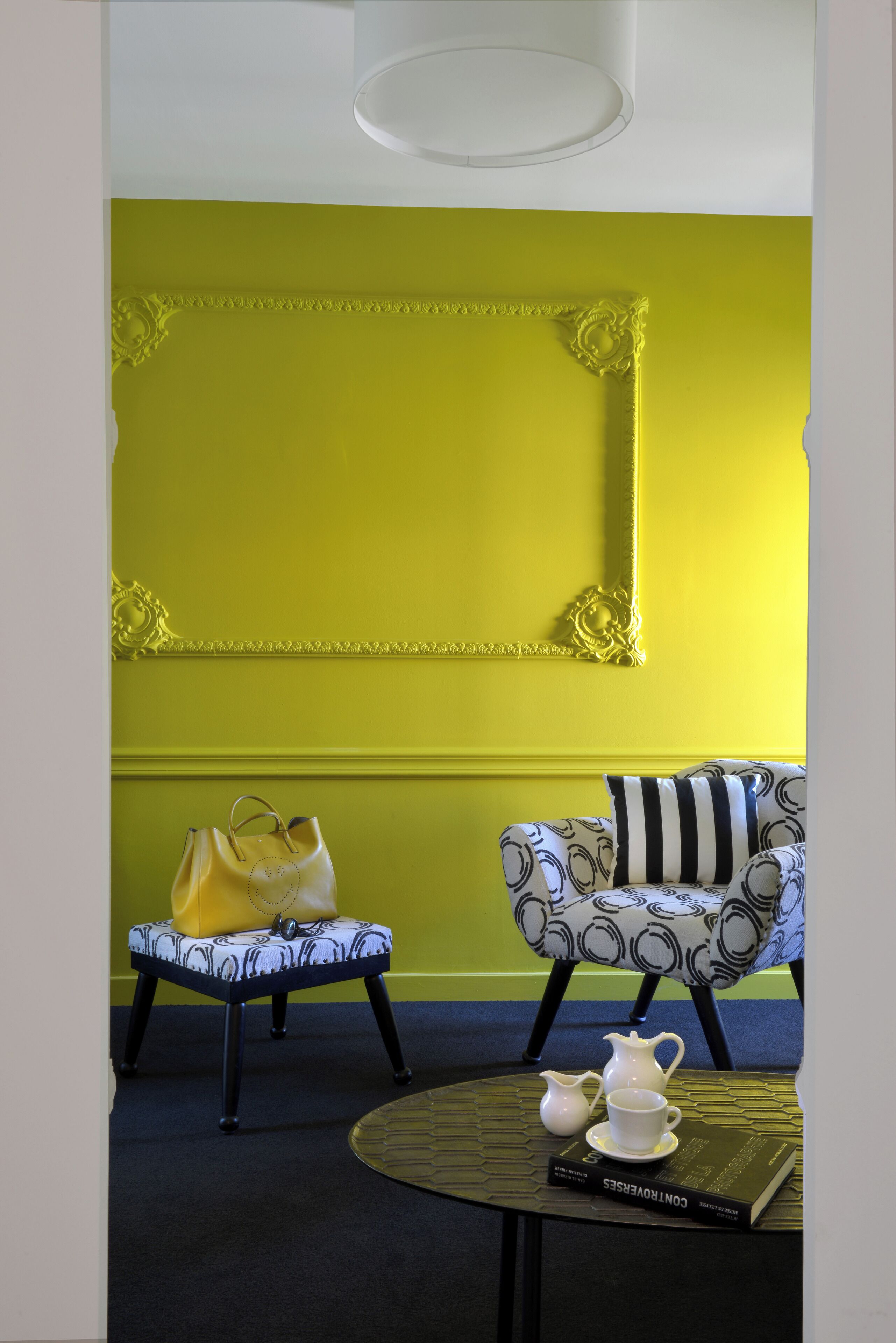 Una habitación con una pared de color amarillo brillante, un sillón estampado, una mesita con un libro y una tetera, y un bolso amarillo sobre un banco.