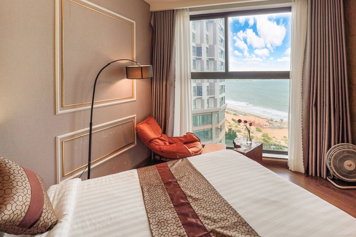 Een hotelkamer met een bed, een stoel, een lamp, een ventilator en uitzicht op de oceaan en gebouwen.