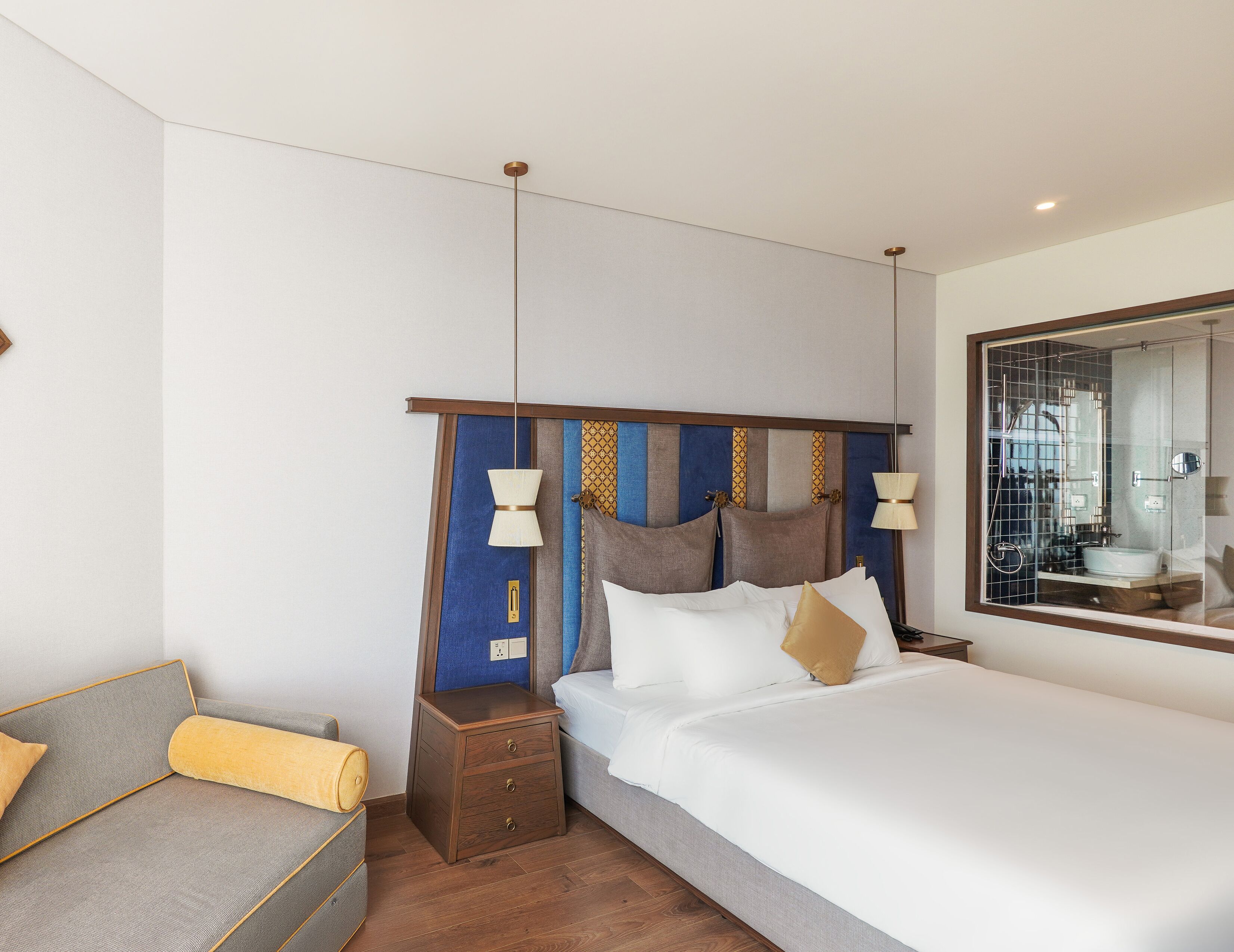 Une chambre d’hôtel moderne avec un grand lit, une table de chevet, un petit canapé et une salle de bain visible à travers une paroi en verre.