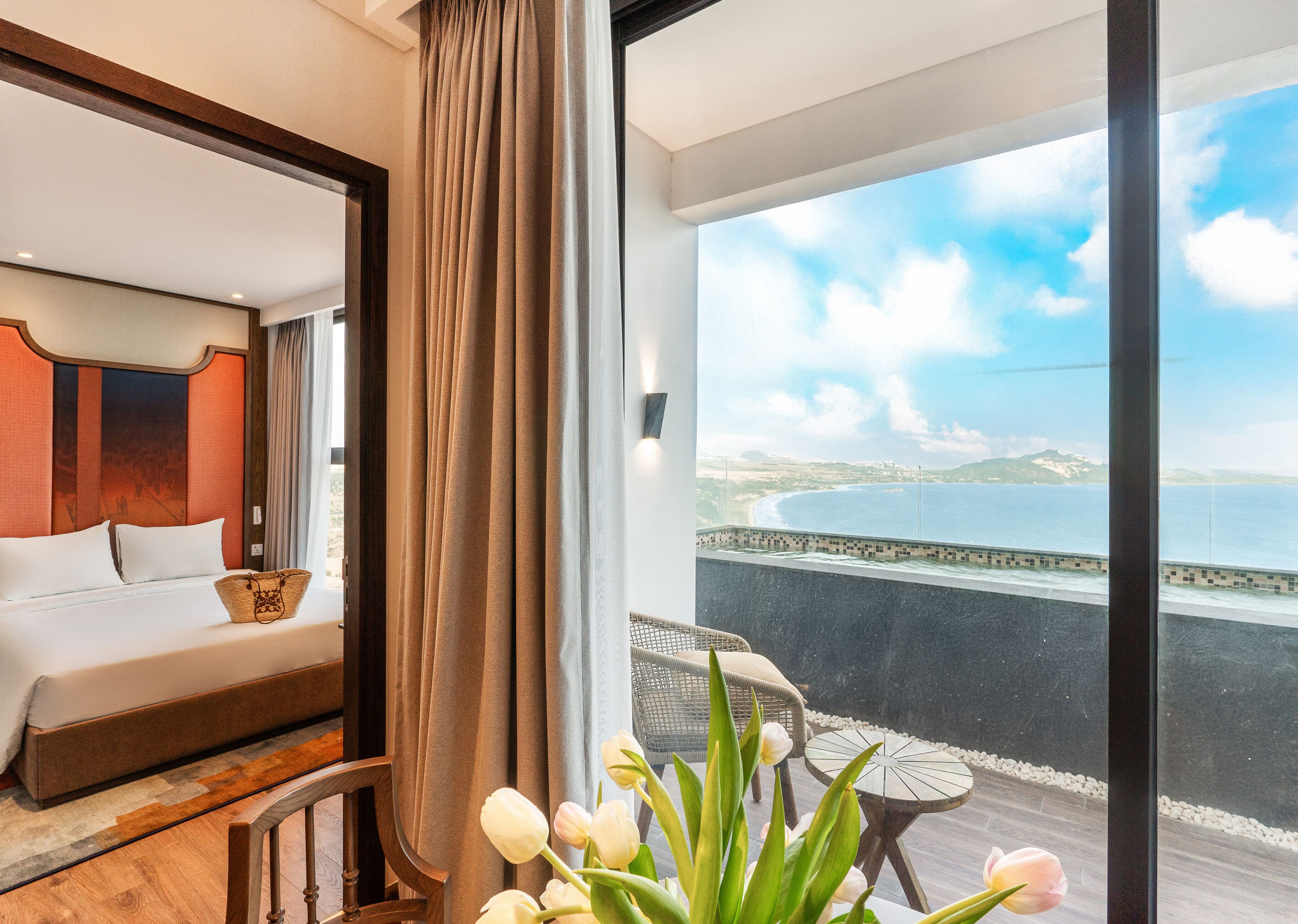 A hotel room with a bed, a view of the sea, and a balcony with a table and chairs.