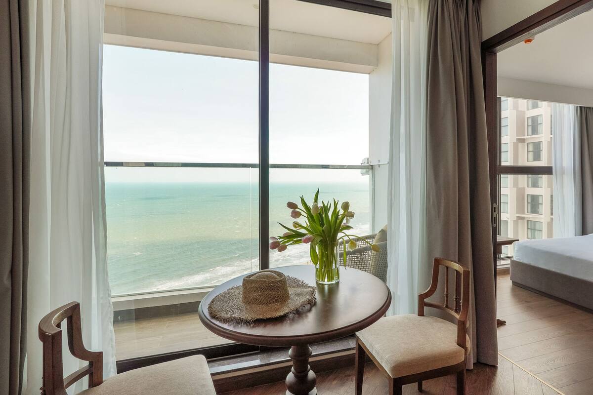 Een hotelkamer met een groot raam, een ronde tafel met een hoed, een stoel, een kleine plant en een bed.