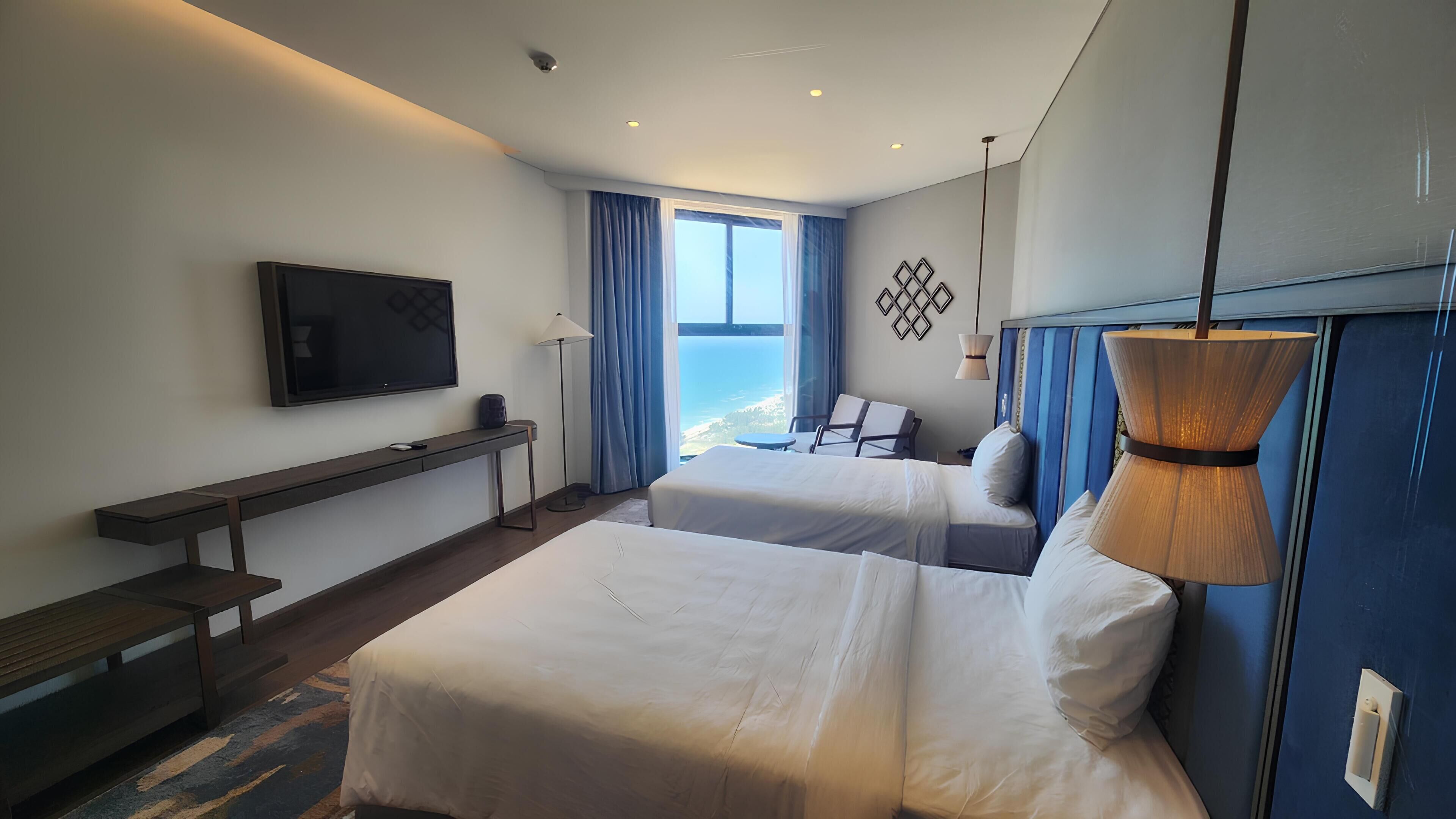 A hotel room with two beds, a TV, a desk, and a view of the sea.