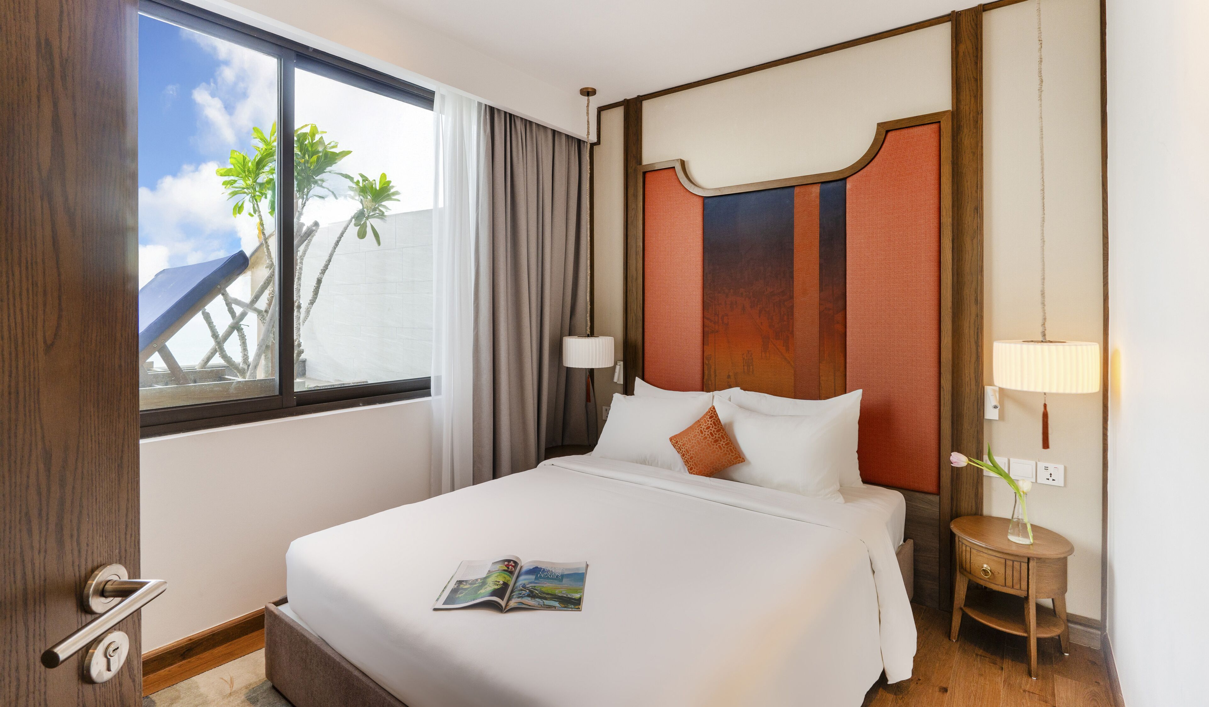 A hotel room with a large bed, a window with a view of palm trees, a wooden headboard, a bedside table with a lamp, and a door with a key in the lock.