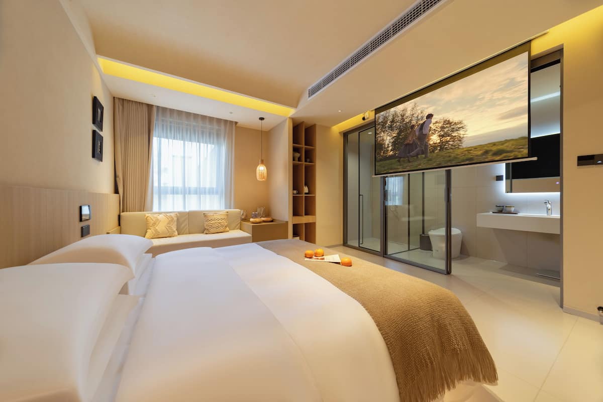 Moderni hotellihuone, jossa on suuri sänky, lasiseinäinen suihkutila ja näkymä kauniille maisemalle.