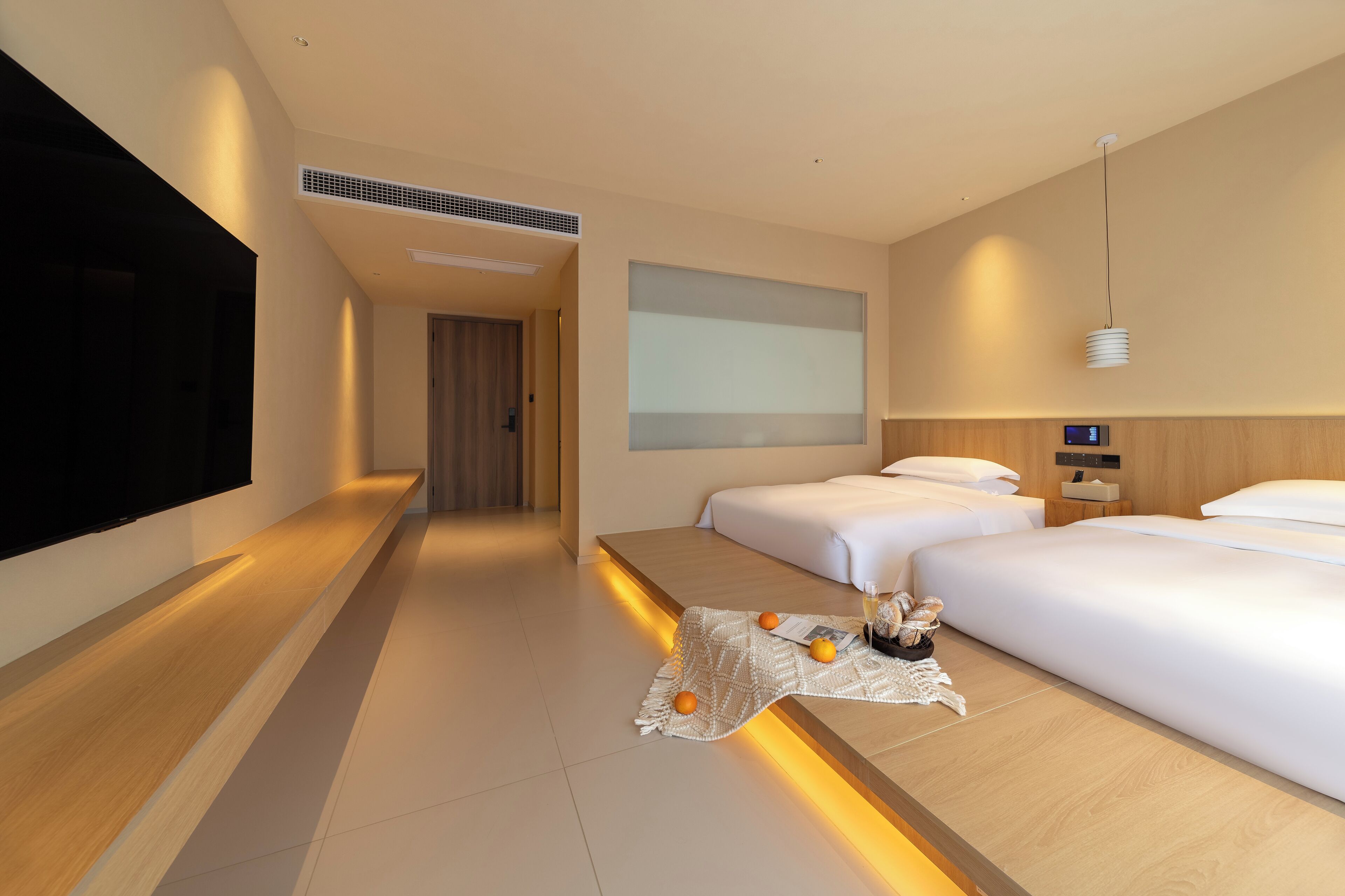 A modern hotel room with a long wooden bench, two beds, a flat-screen TV, and a small table with oranges and bread.