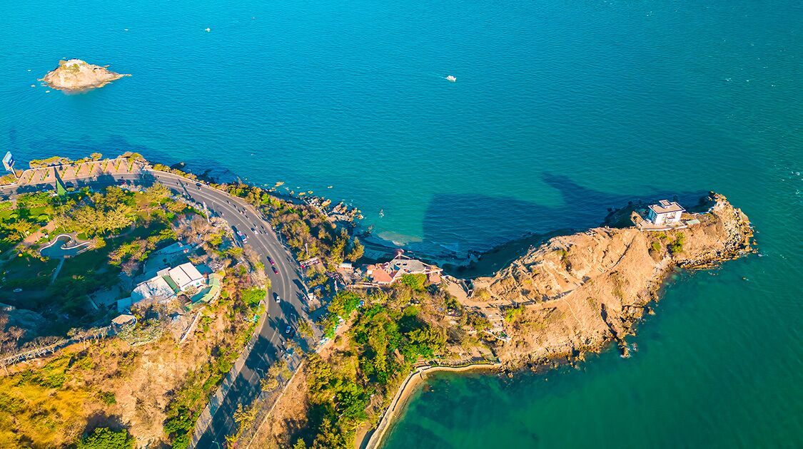 An aerial view of a coastal area with a road curving around a rocky peninsula, a small island, and a building on the peninsula.