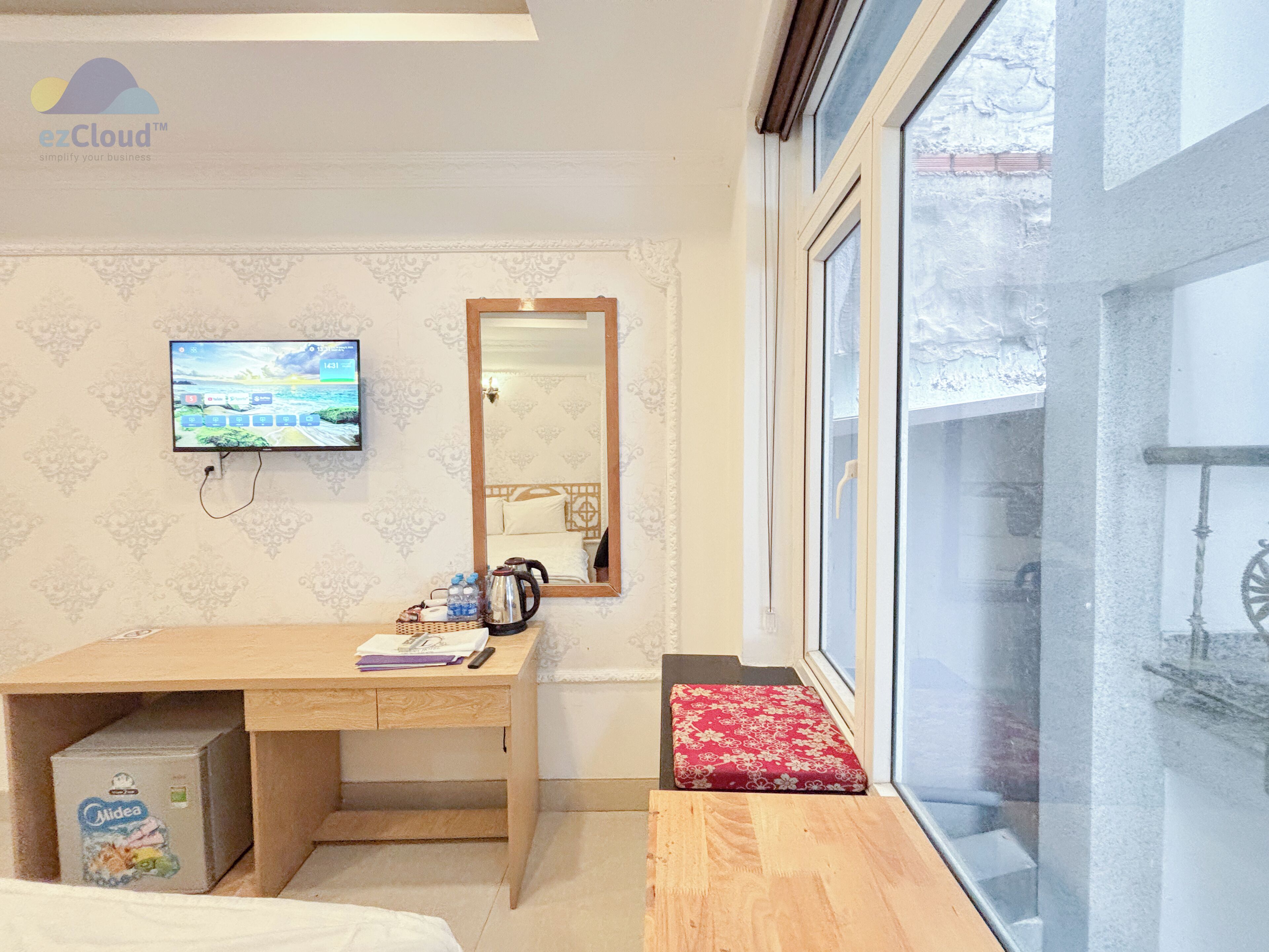 A hotel room with a wooden desk, a television mounted on the wall, a mirror, and a window with a view of the outside.