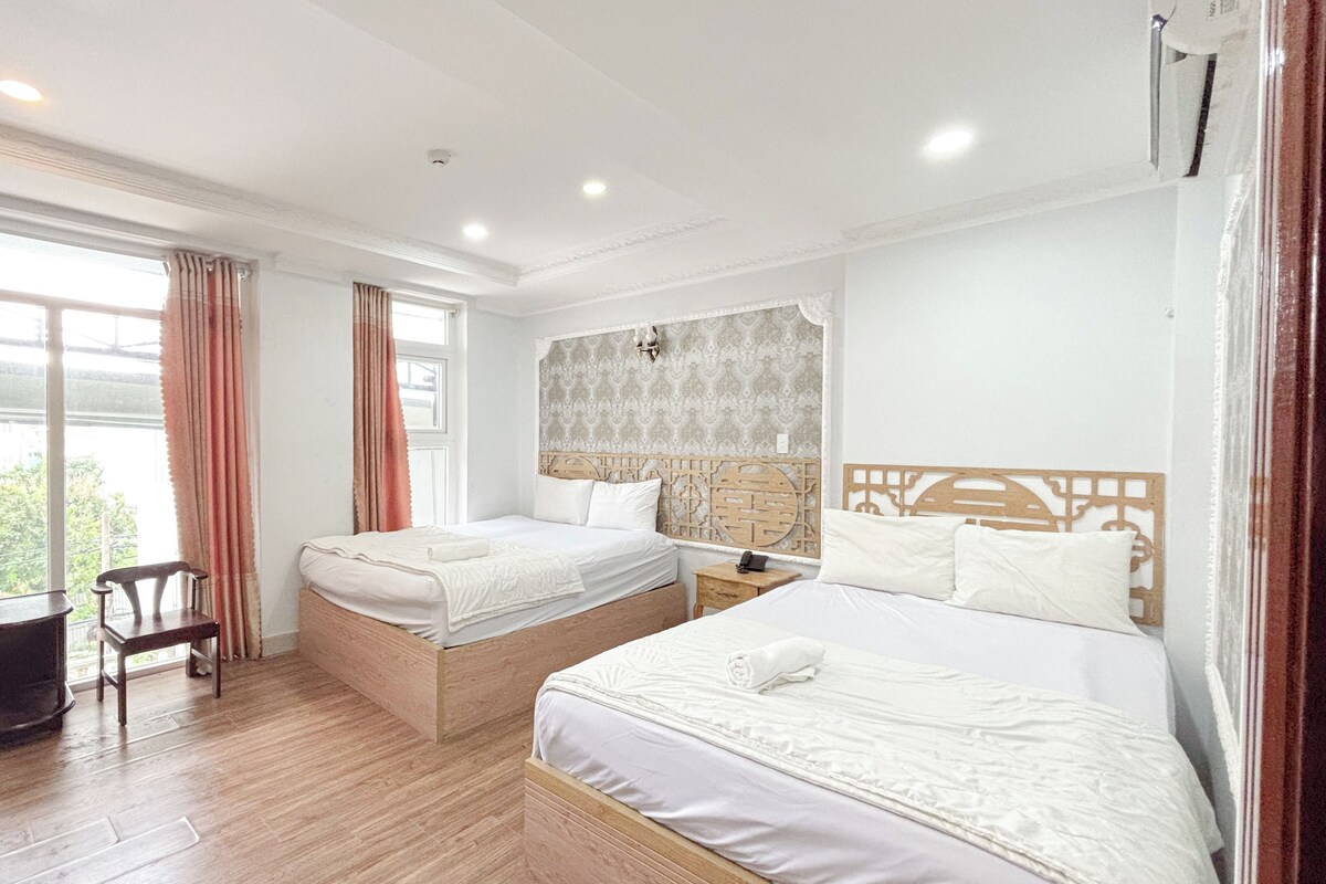 Habitación de hotel con dos camas, suelo de madera, ventana con cortinas y una pequeña mesa con una silla.