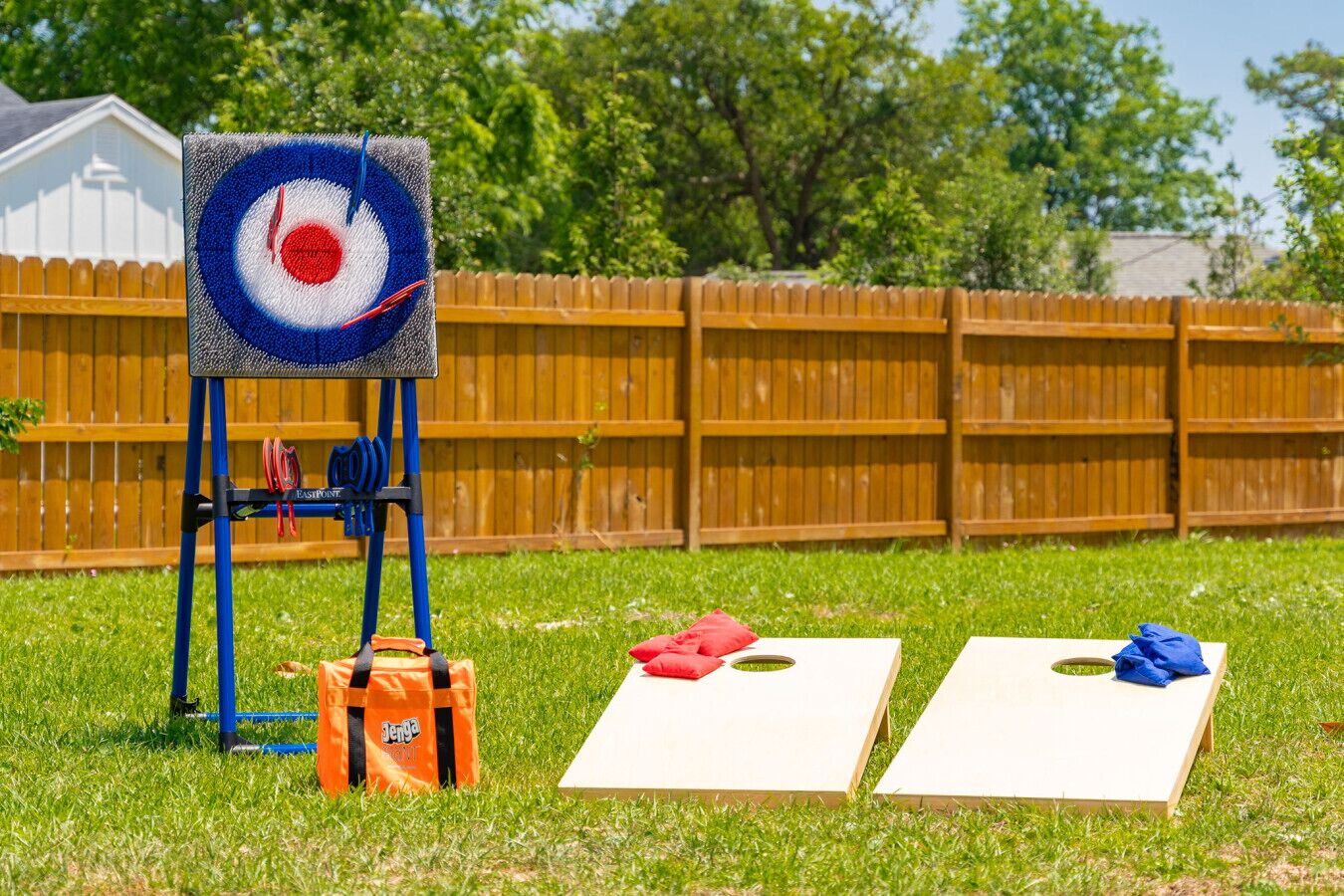 Un jardin avec une cible montée sur un support, deux tapis percés de trous et un sac de jeux Jenga posé sur l’herbe.
