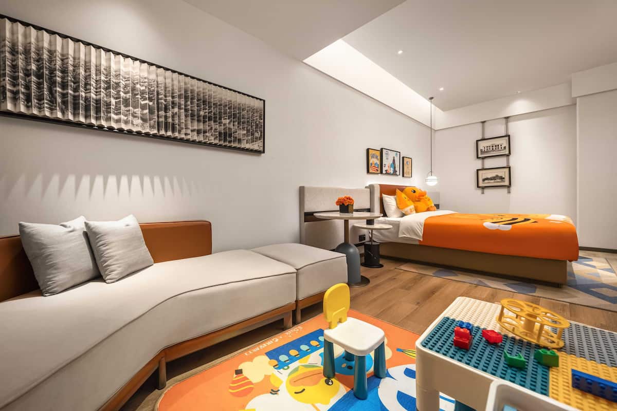 Ett modernt hotellrum med en soffa, en säng med orange sängkläder, ett litet bord med en lampa och en färgglad lekmatta.