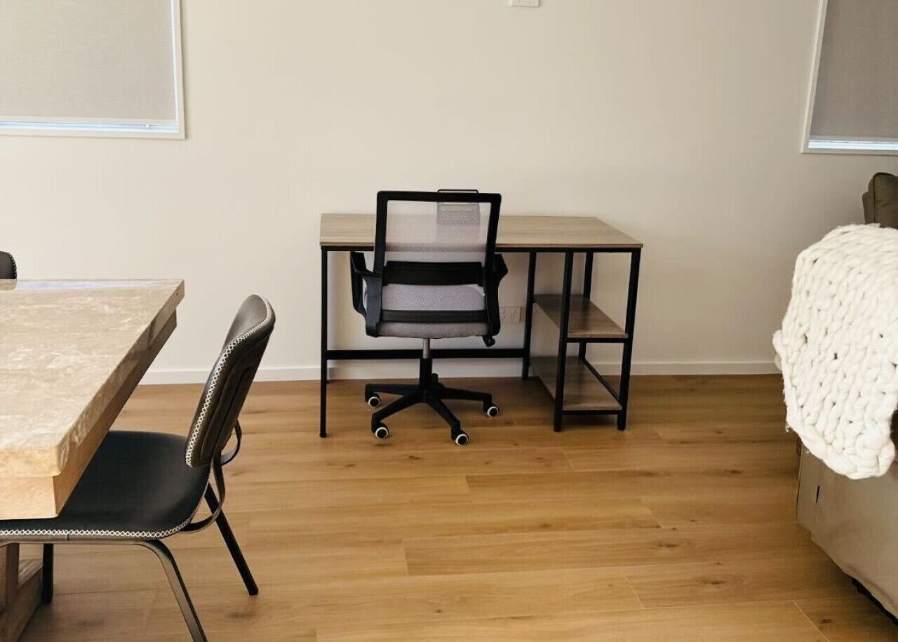 Una habitación con una mesa de comedor de madera, una silla de oficina negra, un escritorio con una silla y una manta blanca de punto en un sofá.