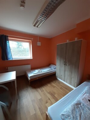 Une petite pièce lumineuse aux murs orange, dotée d’un plancher en bois, d’un lit simple, d’un bureau et d’une armoire.